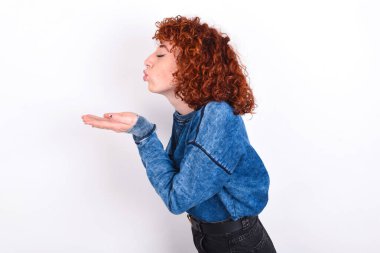 Profile side view view portrait of attractive young caucasian woman red haired wearing blue T-shirt over white background sending air kiss