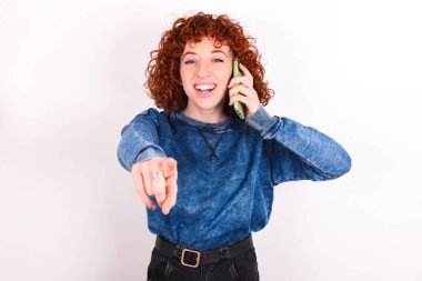 Positive young caucasian woman red haired wearing blue T-shirt over white background indicates directly at camera has telephone conversation smiles broadly enjoys talking long hours. You join me