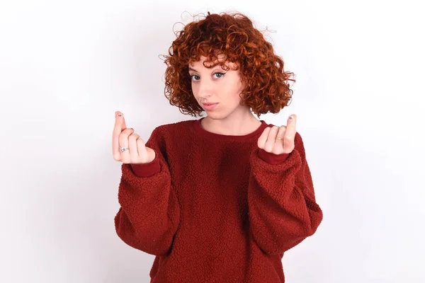 young caucasian woman red haired wearing red sweater over white background keeps doing money gesture with hands, asking for salary payment, millionaire business