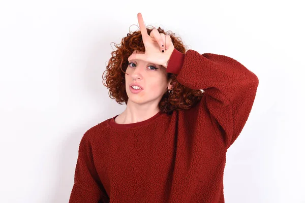 young caucasian woman red haired wearing red sweater over white background keeps making fun of people with fingers on forehead doing loser gesture mocking and insulting.