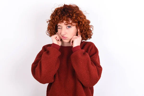 young caucasian woman red haired wearing red sweater over white background keeps covering ears with fingers with annoyed expression for the noise of loud music. Deaf concept.
