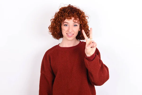 young caucasian woman red haired wearing red sweater over white background keeps showing and pointing up with fingers number two while smiling confident and happy.