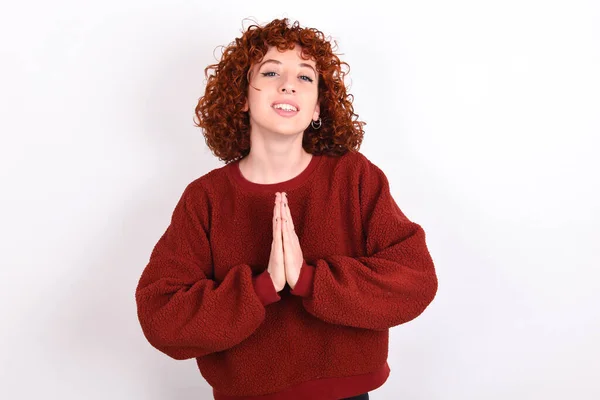 young caucasian woman red haired wearing red sweater over white background keeps praying with hands together asking for forgiveness smiling confident.