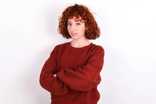 young caucasian woman red haired wearing red sweater over white background keeps happy face smiling with crossed arms looking at the camera. Positive person.