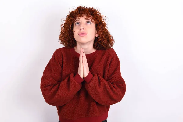 young caucasian woman red haired wearing red sweater over white background keeps begging and praying with hands together with hope expression on face very emotional and worried. Please God