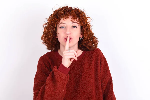 young caucasian woman red haired wearing red sweater over white background keeps makes silence gesture, keeps finger over lips. Silence and secret concept.