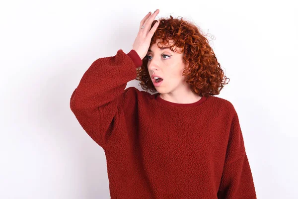 young caucasian woman red haired wearing red sweater over white background keeps surprised with hand on head for mistake, remember error. Forgot, bad memory concept.
