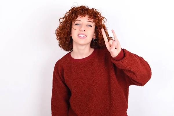 young caucasian woman red haired wearing red sweater over white background keeps doing a rock gesture and smiling to the camera. Ready to go to her favorite band concert.