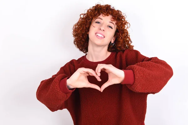 young caucasian woman red haired wearing red sweater over white background keeps smiling in love doing heart symbol shape with hands. Romantic concept.