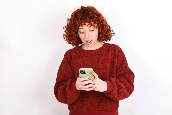 young caucasian woman red haired wearing red sweater over white background keeps using mobile phone chatting free time .