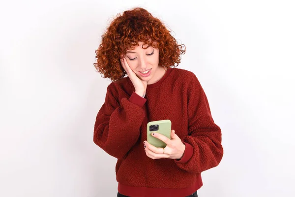 young caucasian woman red haired wearing red sweater over white background keeps holding in hands cell reading sms using new app 5g
