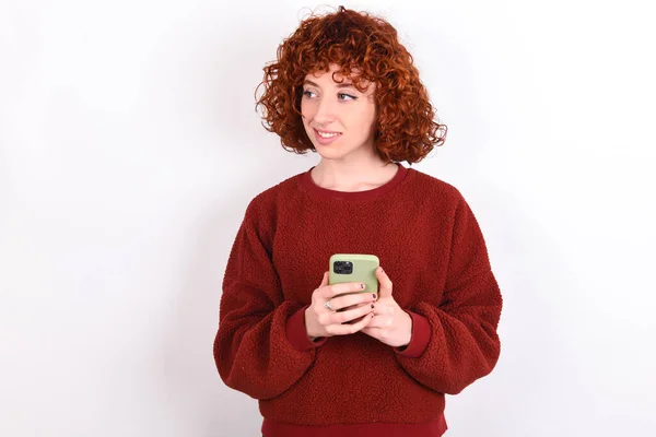 young caucasian woman red haired wearing red sweater over white background keeps hold telephone hands read good youth news look empty space advert