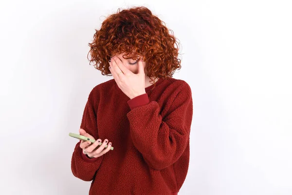 young caucasian woman red haired wearing red sweater over white background keeps looking at smart phone feeling sad holding hand on face.