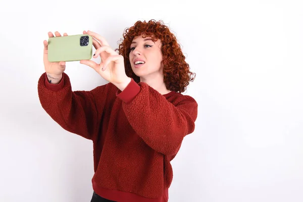 young caucasian woman red haired wearing red sweater over white background keeps taking a selfie to post it on social media or having a video call with friends.