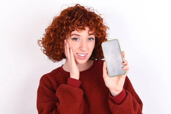 young caucasian woman red haired wearing red sweater over white background keeps hold hand modern technology use touch face palm astonished impressed scream wow omg unbelievable unexpected