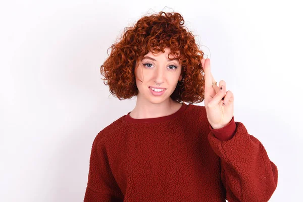 young caucasian woman red haired wearing red sweater over white background keeps pointing up with fingers number ten in Chinese sign language Shi