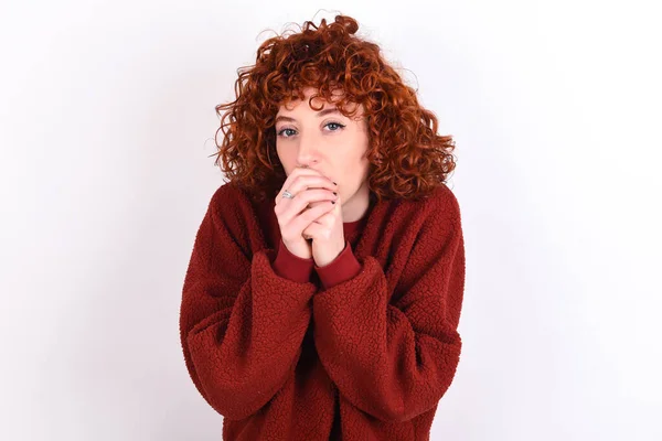 young caucasian woman red haired wearing red sweater over white background keeps holding oneself, feels very cold outside, hopes that will not get cold