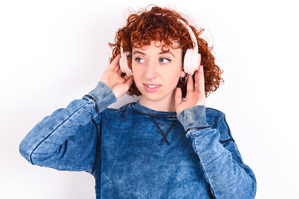 young caucasian woman red haired wearing blue T-shirt over white background wears stereo headphones listens music concentrated aside. People hobby lifestyle concept