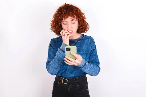 Portrait of pretty frightened young caucasian woman red haired wearing blue T-shirt over white background chatting biting nails after reading some scary news on her smartphone.