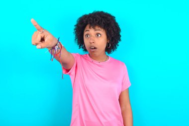 Mavi arka planda pembe tişört giyen genç Afro-Amerikalı kadın. Parmakları önde, ağzı açık, şaşırmış, ön tarafta bir şey var..