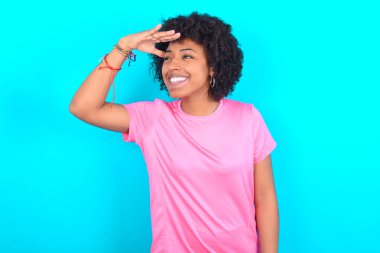 Young African American woman wearing pink T-shirt over blue background very happy and smiling looking far away with hand over head. Searching concept.