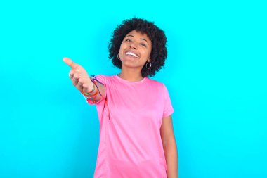 Young African American woman wearing pink T-shirt over blue background smiling friendly offering handshake as greeting and welcoming. Successful business.