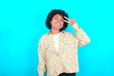 Young African American woman wearing floral shirt over blue background Doing peace symbol with fingers over face, smiling cheerful showing victory
