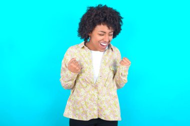 Young African American woman wearing floral shirt over blue background very happy and excited doing winner gesture with arms raised, smiling and screaming for success. Celebration concept.