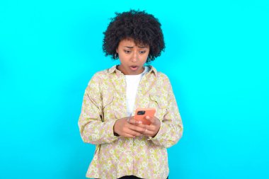 Focused Young African American woman wearing floral shirt over blue background use smartphone reading social media news, or important e-mail