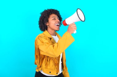 Young African American woman wearing yellow fringe jacket over blue background Through Megaphone with Available Copy Space