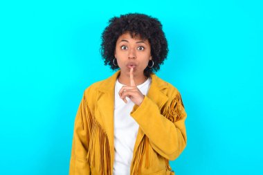 Mavi arka planda sarı püsküllü ceket giyen Afro-Amerikalı kadın sessiz bir jest yapıyor, parmaklarını dudaklarının üstünde tutuyor ve kamerada gizemli görünüyor.