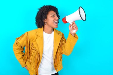 Mavi arka planda sarı püsküllü ceket giyen komik, genç Afrikalı Amerikalı kadın. Kopyalama alanı oluşturun. Megafonla bağırıyordu..