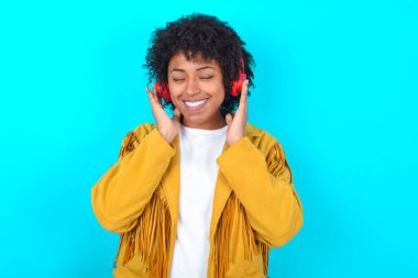 Mavi arka planda sarı püsküllü ceket giyen memnun genç Afrikalı Amerikalı kadın hoş melodiyi dinlemekten zevk alıyor. Müzik seti kulaklığı takıyor, gözleri kapatıyor. Müzikle boş zaman geçirmek