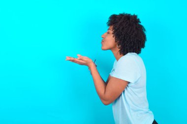 Afro saçlı, mavi arka planda mavi tişört giyen, hava öpücüğü gönderen çekici Afro-Amerikan kadın portresi.