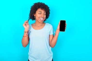 Mavi arka planda mavi tişört giyen güzel bir Afro-Amerikan kadının fotoğrafı telefon ekranı saç kuyruğu olduğunu gösteriyor.