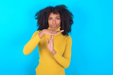 Mavi arka plan üzerinde sarı boğazlı kazak giyen Afro-Amerikalı bir kadın üzgün, mola verme hareketi gösteriyor, durması gerekiyor, çok çalıştıktan sonra dinlenmesi gerekiyor, el işaretini kırdığını gösteriyor