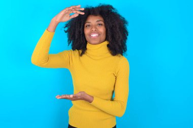 Mavi arka planda sarı boğazlı kazak giyen Afro-Amerikalı kadın. Elleri büyük ve büyük boylarda işaret ediyor. Ölçüm sembolü. Kameraya bakıp gülümsüyor..