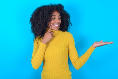 Mavi arka plan üzerine sarı balıkçı yaka giyen komik Afro-Amerikan kadın elinde yeni bir şey tutuyor. Satın almak istiyorum.!