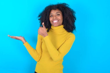 Mavi arka planda sarı boğazlı kazak giyen Afro-Amerikalı kadın avuç içini gösteriyor ve baş parmaklarıyla hareket ediyor, mutlu ve neşeli gülümsüyor..