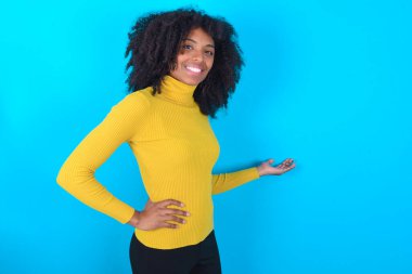 Mavi arka planda sarı boğazlı kazak giyen Afro-Amerikalı kadın mutlu ve neşeli hissediyor, size gülümsüyor ve hoş geldiniz diyor, dostça bir jestle sizi içeri davet ediyor.