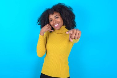 Mavi arka planda sarı boğazlı kazak giyen Afro-Amerikalı kadın neşeli bir şekilde gülümsüyor ve kamerayı işaret ediyor. Daha sonra seni arıyor, telefonla konuşuyor.