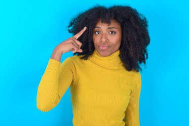 Mavi arka plan üzerinde sarı boğazlı kazak giyen Afro-Amerikalı kadın bir şey hatırlamaya çalışıyor, parmağını şakağa uzatıyor, sınav için gerekli bilgileri hatırlatıyor.,