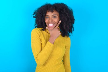 Mavi arka planda sarı boğazlı kazak giyen Afro-Amerikalı kadın çapraz kollu ve çeneli bir şekilde gülümseyen kameraya kendinden emin bakıyor. Olumlu düşünüyorum..