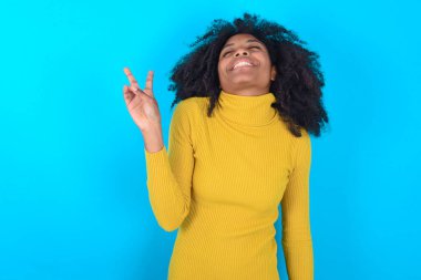 Mavi arka planda sarı boğazlı kazak giyen Afro-Amerikalı kadın kameraya gülümseyen ve zafer işareti yapan. İki numara..