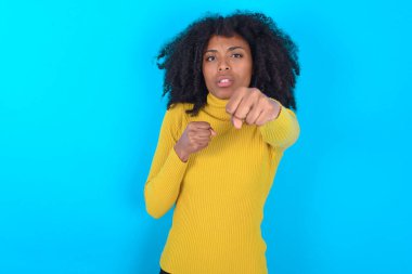 Mavi arka planda sarı boğazlı kazak giyen Afro-Amerikalı kadın dövüşmek için yumruk, agresif ve öfkeli saldırı, tehdit ve şiddet