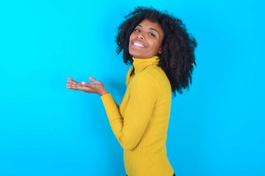 Mavi arka plan üzerine sarı balıkçı yaka giyen Afro-Amerikalı kadın avuçlarını açıp fotokopi alanını gösteriyor.