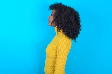 Mavi arka planda sarı boğazlı kazak giyen Afro-Amerikalı kadın, yan tarafa bakıyor, rahat poz veriyor, kendine güvenen bir gülümsemesi var..