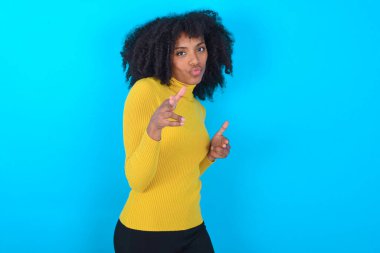 Mavi arka plan üzerinde sarı boğazlı kazak giyen neşeli Afro-Amerikalı kadın kameraya işaret parmağıyla işaret ediyor, birini seçiyor, parmak tabancası yapıyor..