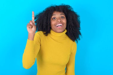 Mavi arka planda sarı boğazlı kazak giyen Afro-Amerikalı kadın parmağıyla işaret ediyor ve dahiyane düşüncelerden esinlenerek bakıyor, iyi bir fikir işareti gösteriyor, zekice bir çözümü var.