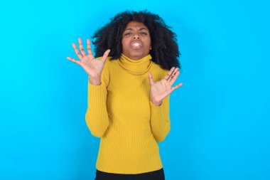 Mavi arka planda sarı boğazlı kazak giyen Afro-Amerikalı kadın korku ifadesinden korkuyor ve dehşete düşüyor. El hareketlerini durdur, şok içinde bağır. Panik konsepti.